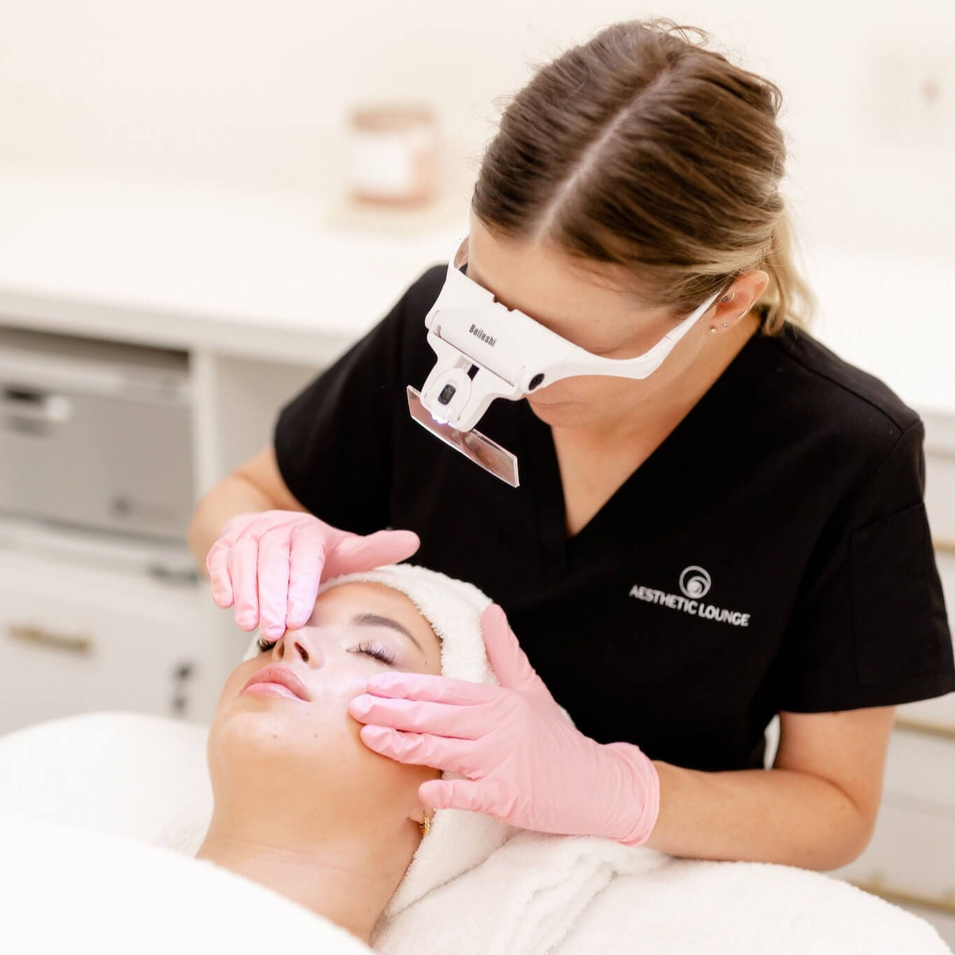 Professional facial treatment being performed by a licensed aesthetician wearing magnifying goggles and pink gloves, applying skincare to a female client's face at Aesthetic Lounge