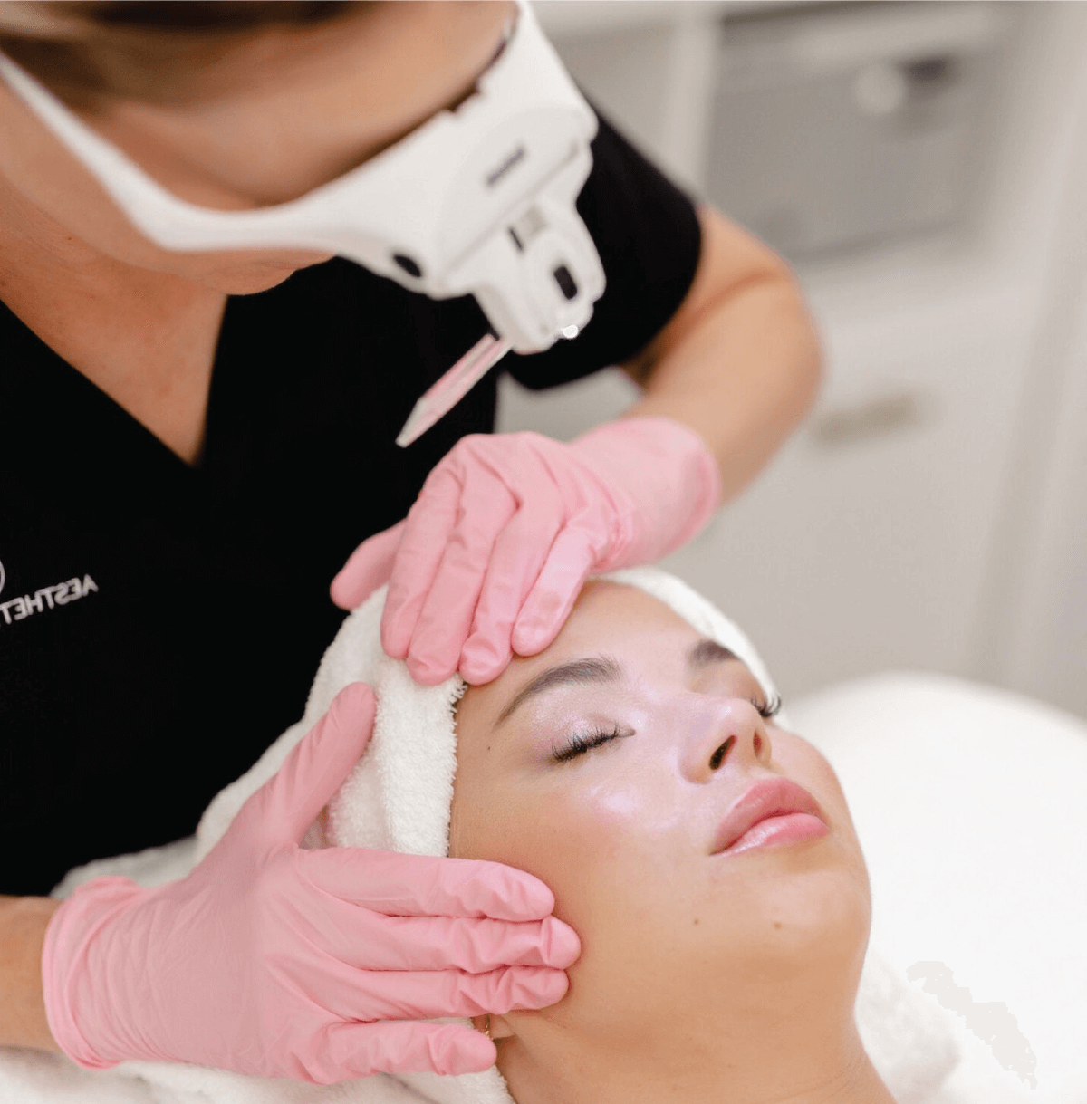 Woman receiving a relaxing facial treatment while wearing a headband, with an aesthetician in black uniform and pink gloves gently massaging her face