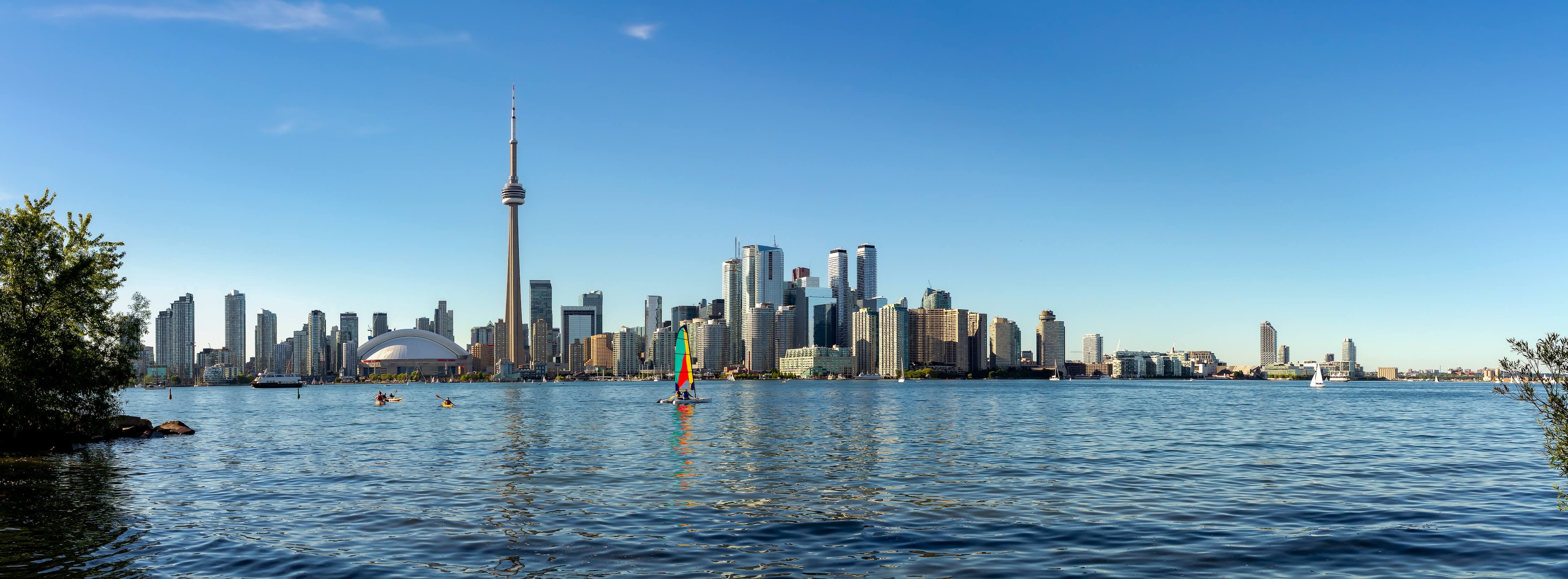 Panoramic view of a city skyline with a prominent tower, reflected in water, under a clear blue sky.
