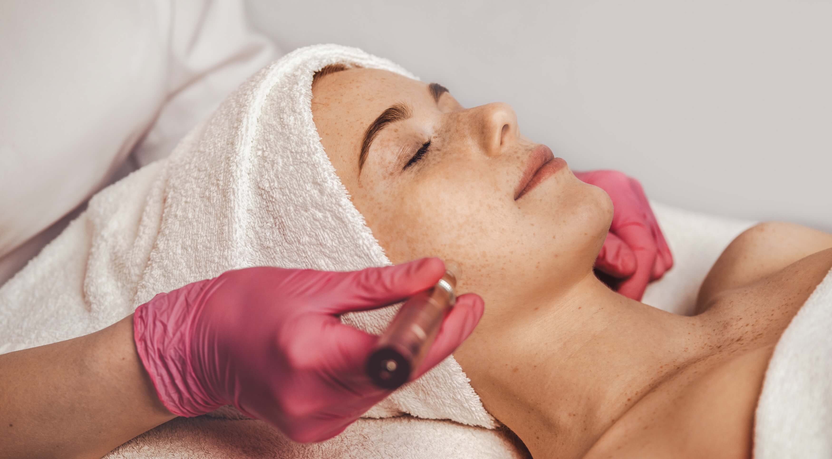 Microneedling procedure being performed on a woman’s face, with technician wearing pink gloves and patient lying relaxed with a towel wrap