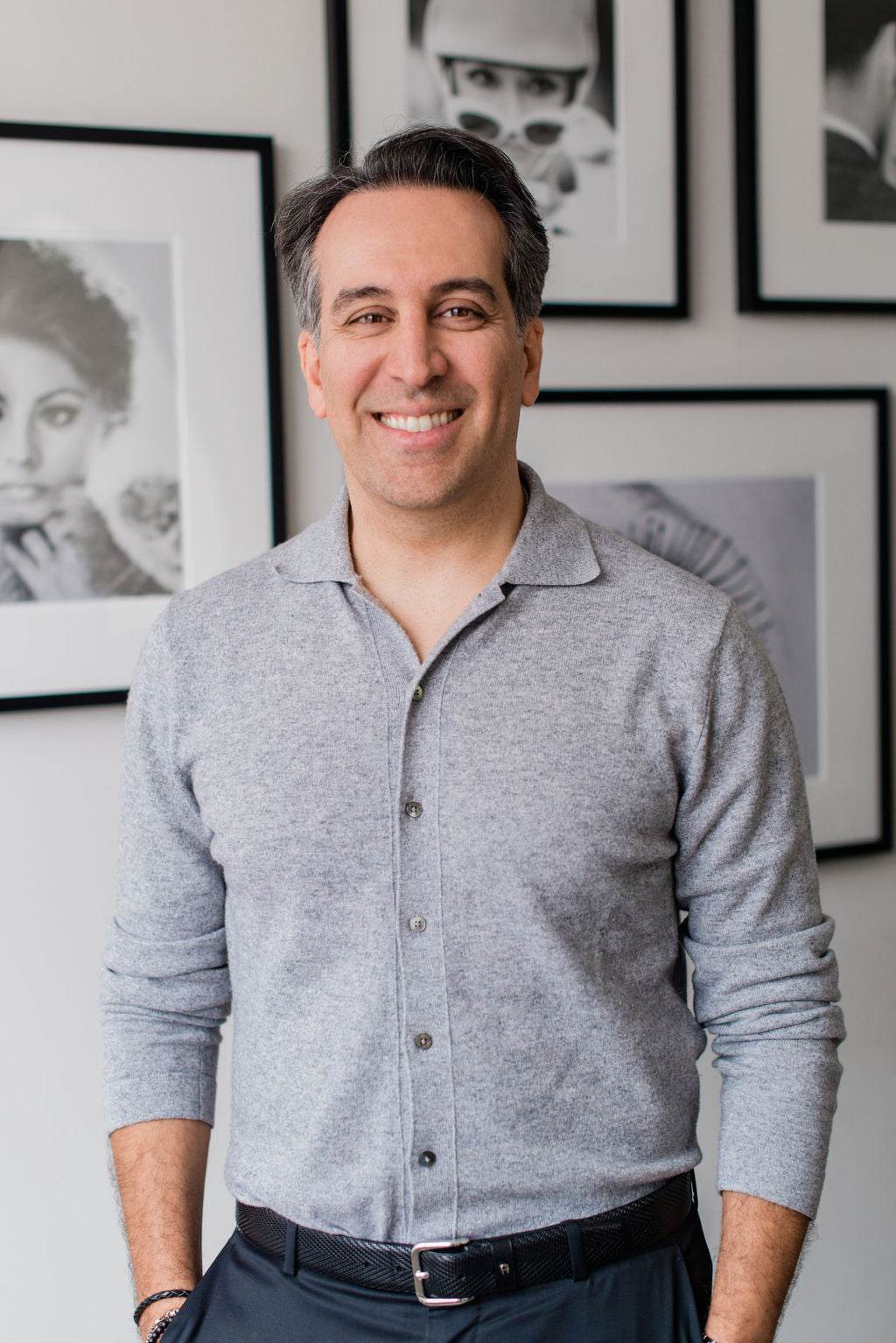 Dr. Babak Maleki smiling in a casual outfit, standing confidently in front of black-and-white portrait wall at his clinic