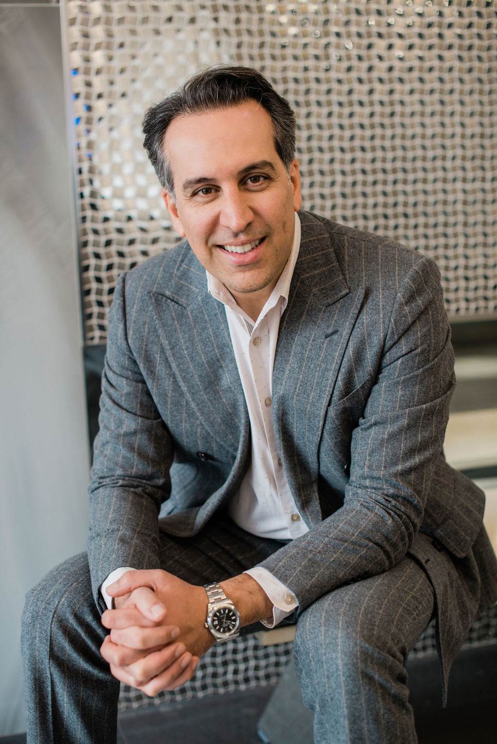 Dr. Babak Maleki wearing a tailored grey pinstripe suit, smiling confidently while seated in an elegant clinic lounge