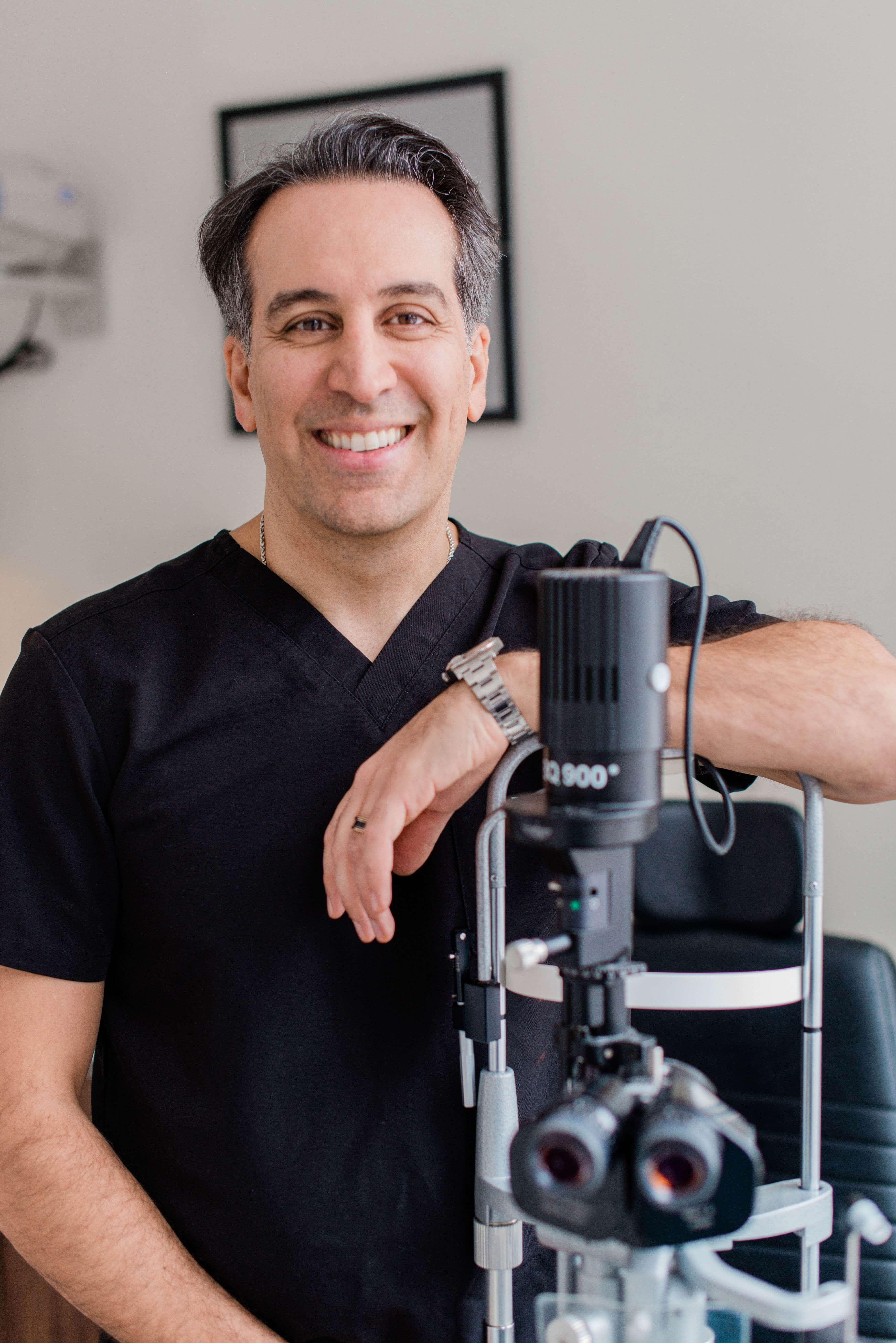 Dr. Babak Maleki smiling in black scrubs, standing beside advanced diagnostic equipment at his cosmetic clinic