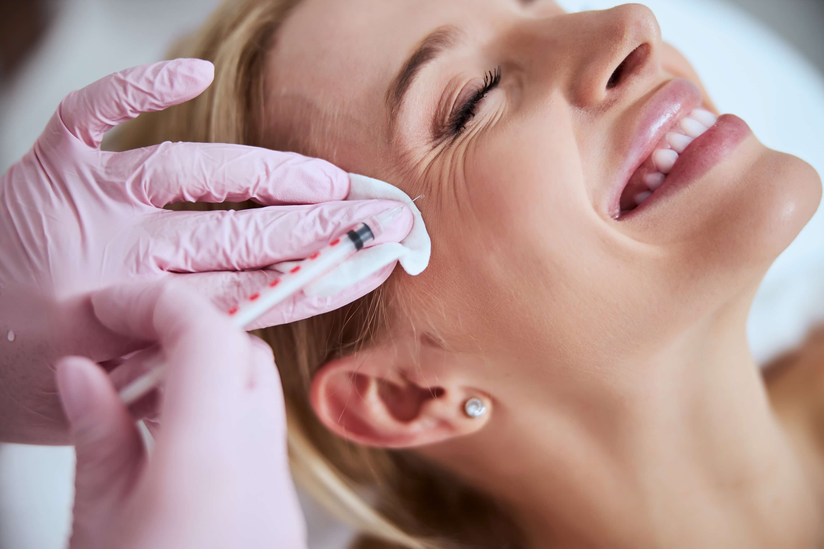 Smiling woman receiving a Botox injection near the eye area, performed by a professional wearing pink gloves in a clean clinical setting