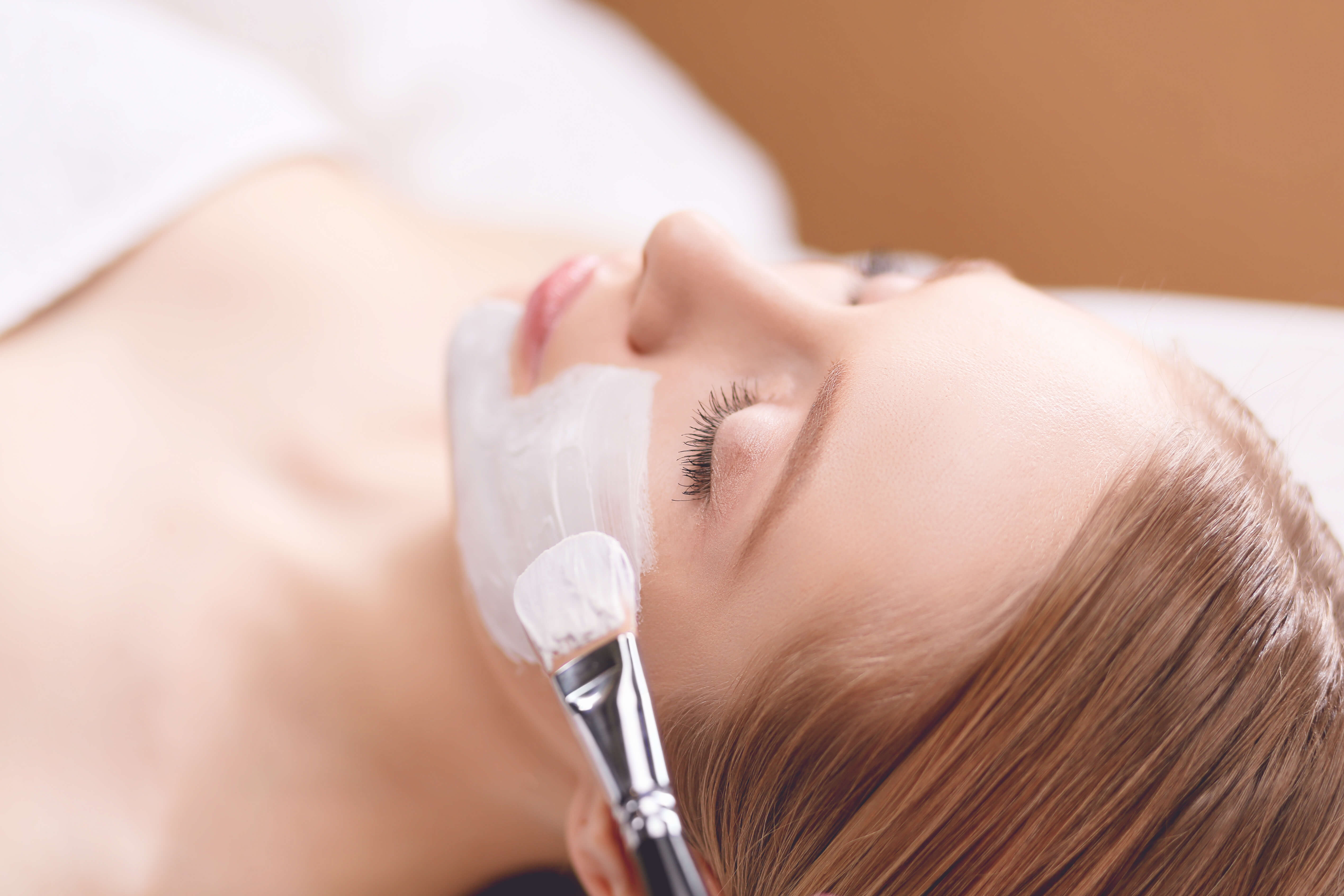 Woman receiving chemical peel treatment with a brush applying white exfoliating solution to the face for skin renewal and texture improvement.