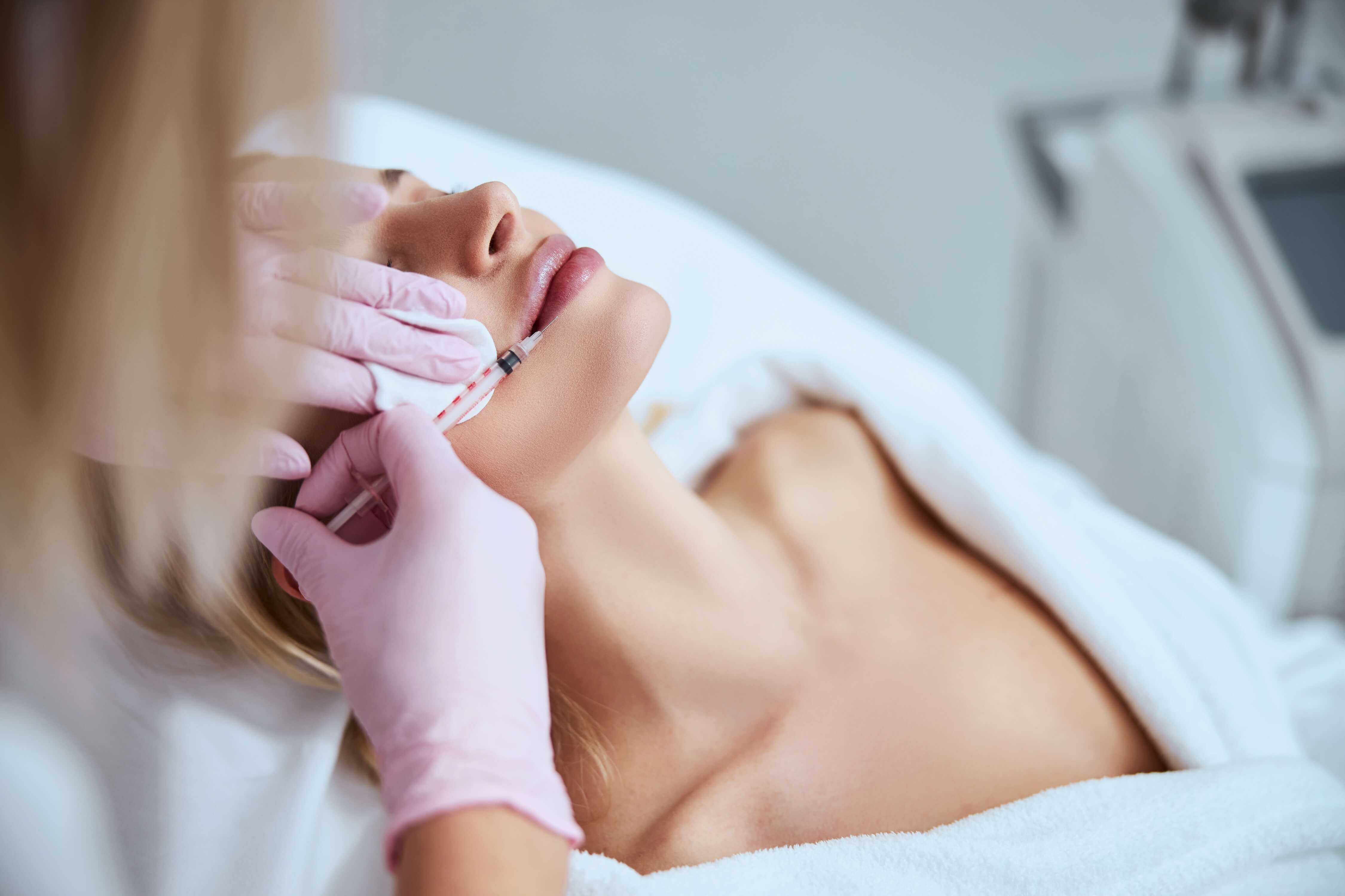 Lip filler injection performed by a professional wearing pink gloves on a woman lying in a clinical treatment room
