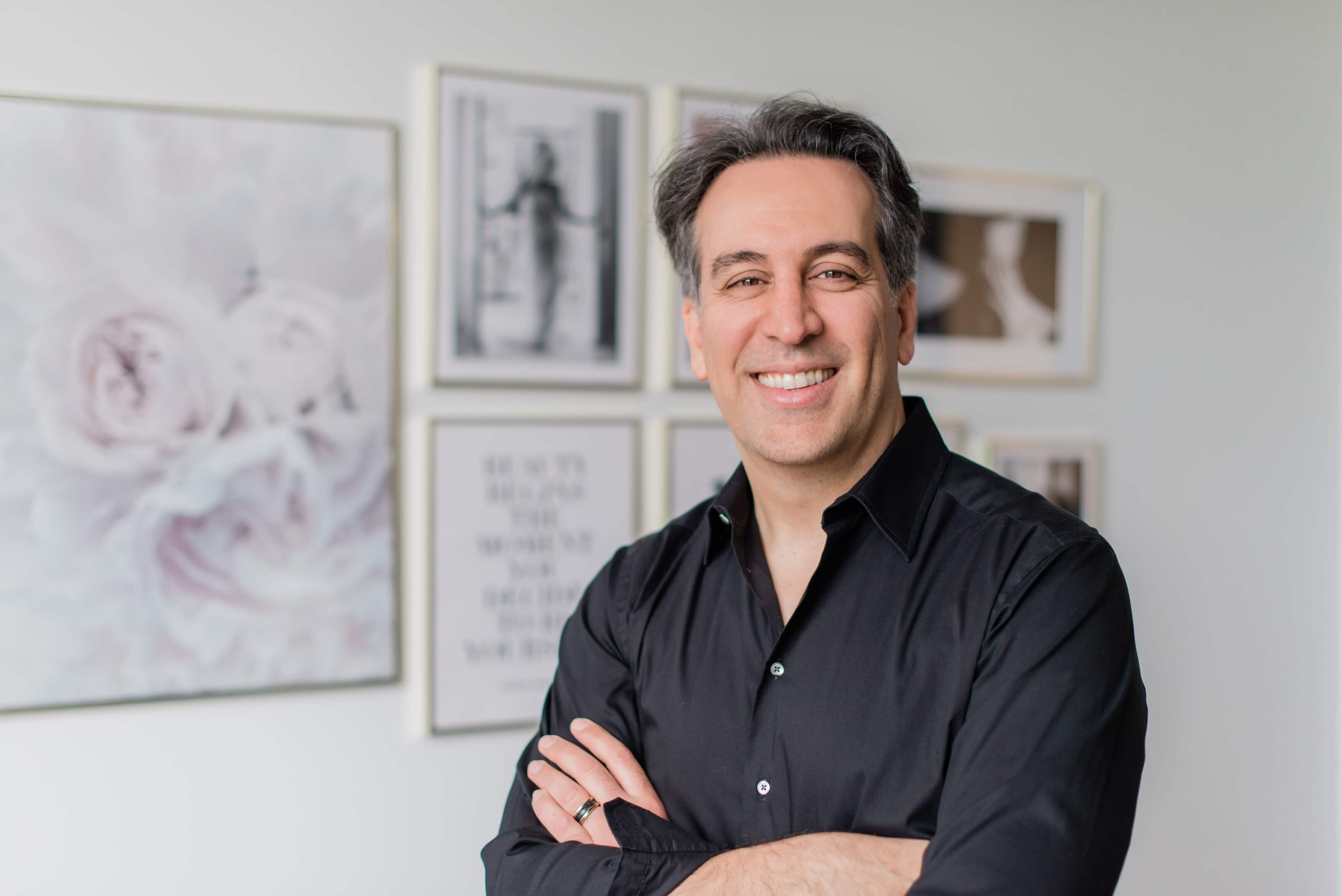 Portrait of Dr. Babak Maleki smiling in a black shirt, standing in front of modern framed artwork at his Toronto clinic