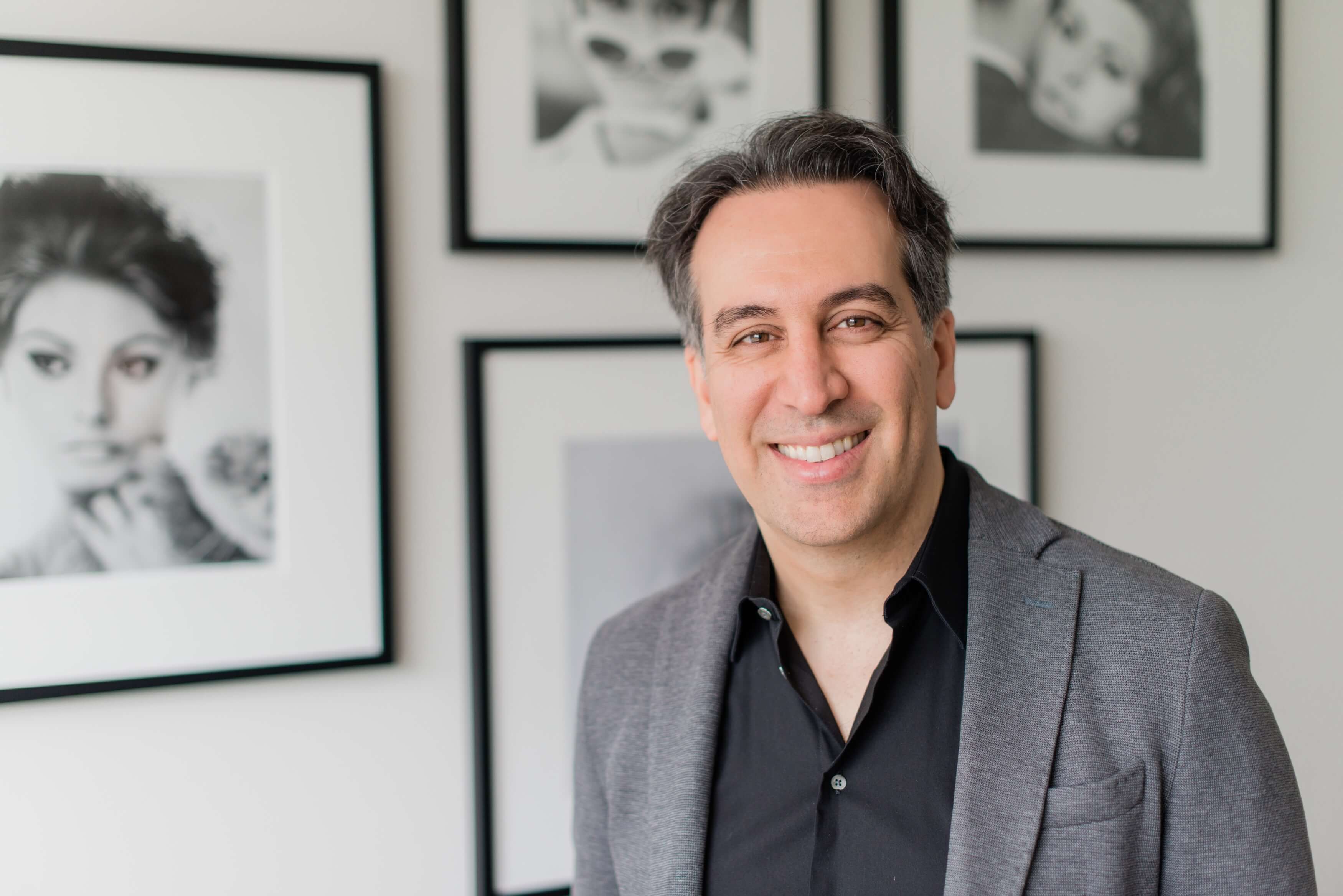 Dr. Babak Maleki smiling in a grey blazer and black shirt, standing in front of black-and-white portrait artwork at his clinic