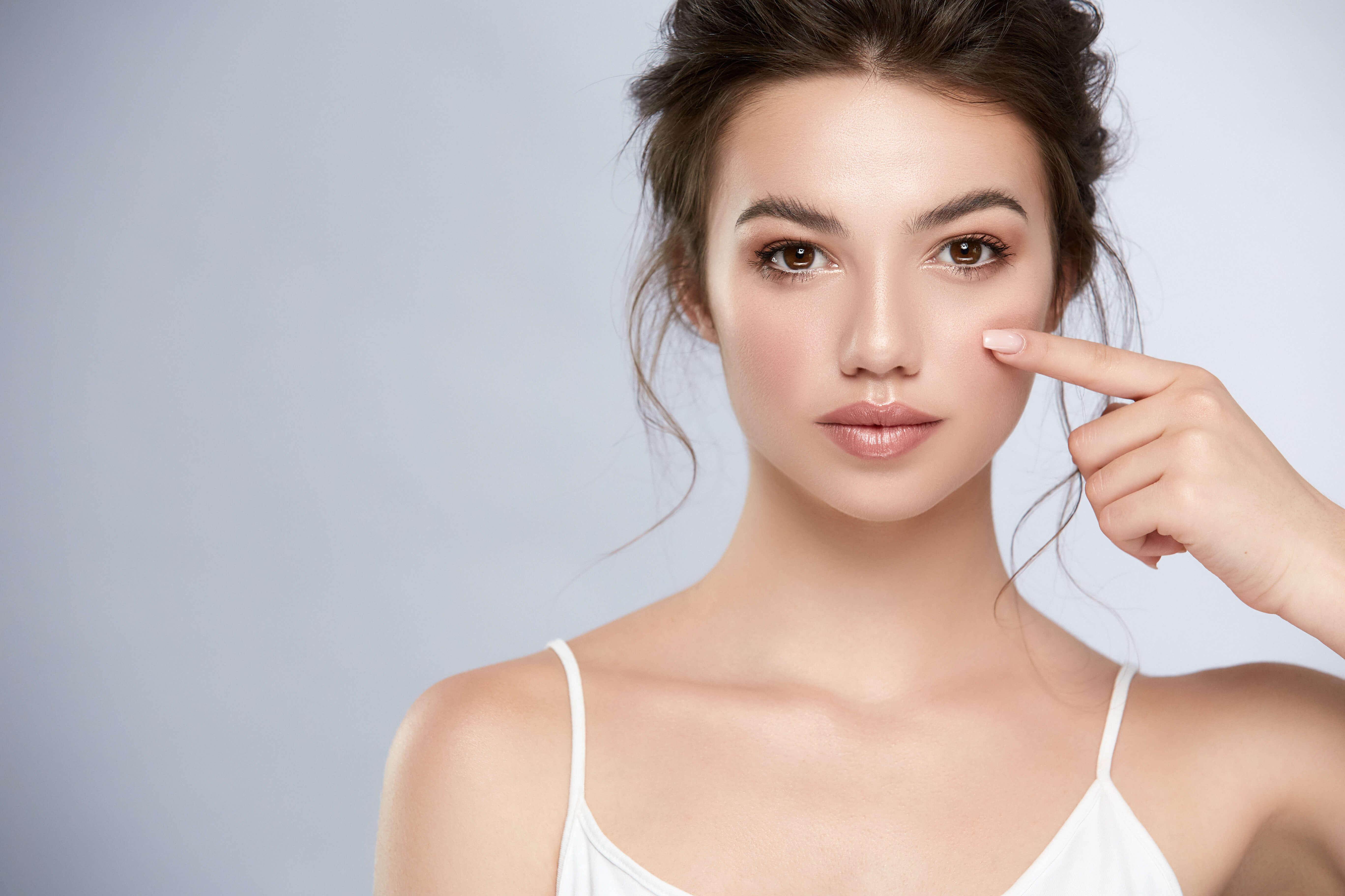 Beautiful young woman with flawless skin pointing to under-eye area, ideal for promoting filler, skincare, or cosmetic treatments at Dr. Babak Maleki’s clinic