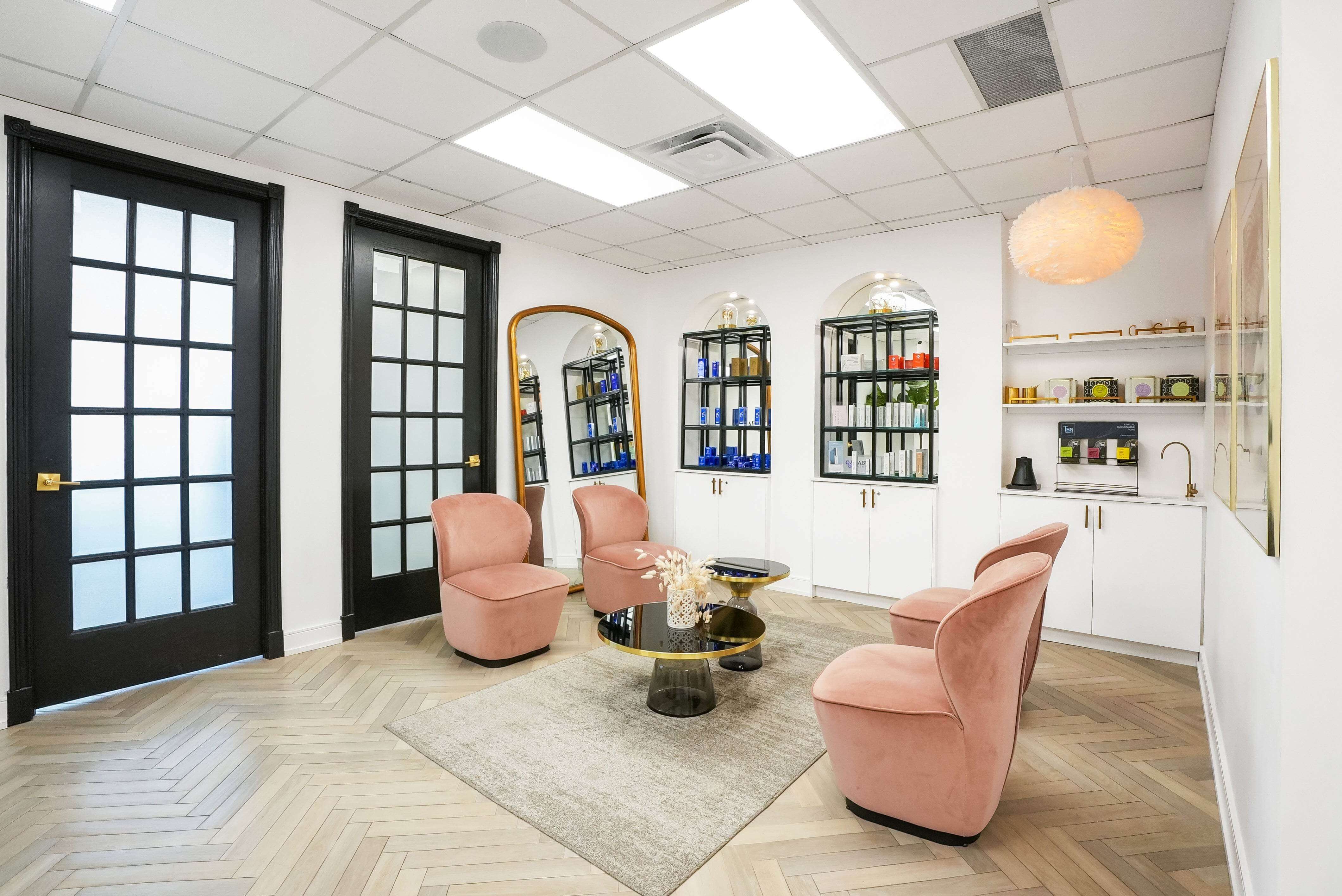 Elegant waiting room with soft blush chairs, black-framed glass doors, and modern skincare display shelves at Aesthetic Lounge by Dr. Maleki